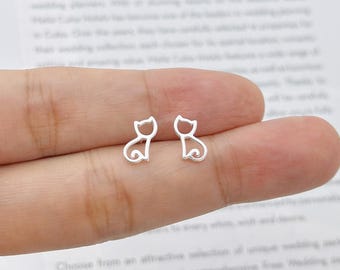 May include: A pair of silver cat-shaped stud earrings. Each earring features a minimalist outline of a cat with a curled tail. The earrings are small and delicate, perfect for everyday wear. The background is a blurred image of text.