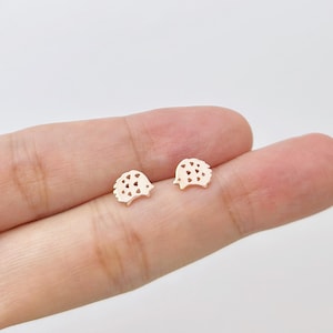 May include: A pair of small, gold-colored hedgehog stud earrings. Each earring features a detailed hedgehog silhouette with cut-out details. The earrings are displayed on a light background, showcasing their delicate design and craftsmanship.