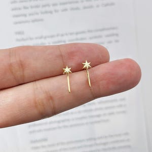 May include: A pair of gold-colored star-shaped stud earrings. Each earring features a five-pointed star atop a slender, straight post. The earrings are displayed against a blurred background, highlighting their delicate design.