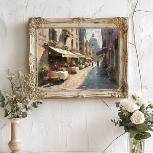 May include: A framed painting of a street scene in Venice, Italy. The painting depicts a narrow street lined with buildings, with a cafe on the left side of the street. The cafe has tables and chairs set up outside, and there are people sitting at the tables. In the background, there is a large dome-shaped building, which is likely the Basilica di San Marco.