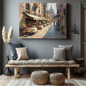 May include: A painting of a street scene in Venice, Italy. The painting shows a narrow street lined with buildings, with a cafe on the left side of the street. The cafe has tables and chairs set up outside, and there are people sitting at the tables. In the background, there is a large dome-shaped building, which is likely the Basilica di San Marco.