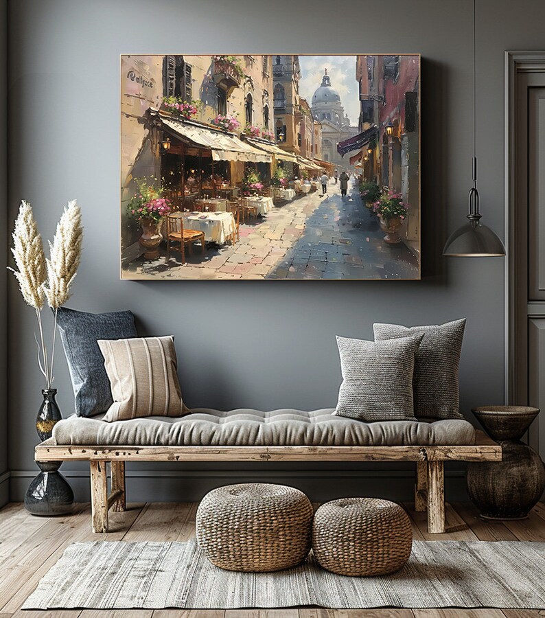 May include: A painting of a street scene in Venice, Italy. The painting shows a narrow street lined with buildings, with a cafe on the left side of the street. The cafe has tables and chairs set up outside, and there are people sitting at the tables. In the background, there is a large dome-shaped building, which is likely the Basilica di San Marco.