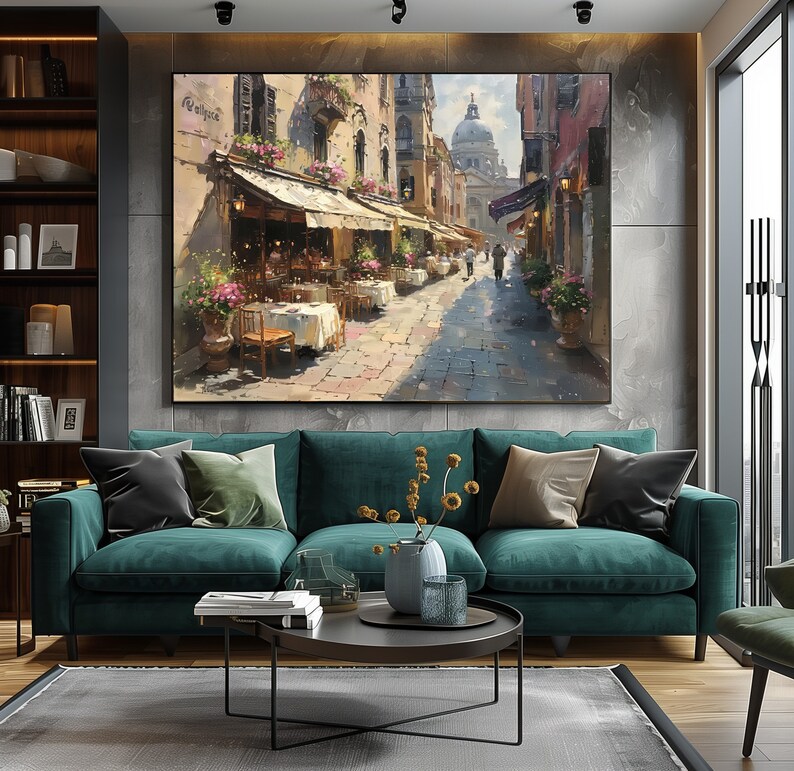 May include: A green velvet sofa with two throw pillows, a coffee table with a vase of dried flowers, and a painting of a street scene in Venice, Italy. The painting features a cafe with tables and chairs, and people walking down the street.