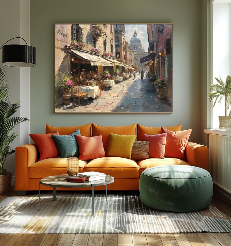May include: A painting of a street scene in a European city. The street is lined with buildings and there are people walking by. There are tables and chairs set up outside of a cafe. The painting is done in a realistic style and has a warm, inviting feel.