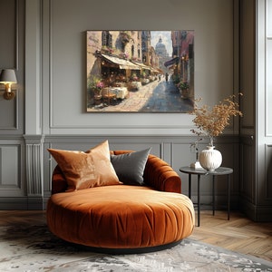 May include: A round, orange velvet chaise lounge with two pillows, one brown and one gray, in a room with gray walls and a painting of a street scene with a building in the background.