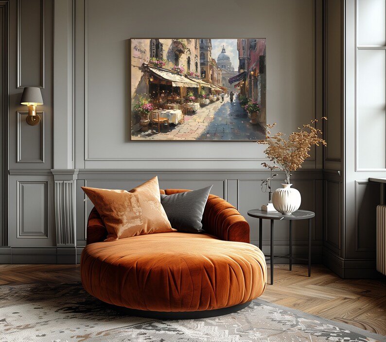 May include: A round, orange velvet chaise lounge with two pillows, one brown and one gray, in a room with gray walls and a painting of a street scene with a building in the background.