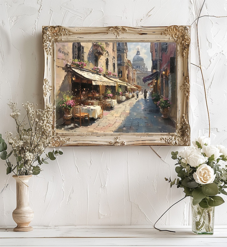 May include: A framed painting of a street scene in Venice, Italy. The painting depicts a narrow street lined with buildings, with a cafe on the left side of the street. The cafe has tables and chairs set up outside, and there are people sitting at the tables. In the background, there is a large dome-shaped building, which is likely the Basilica di San Marco.