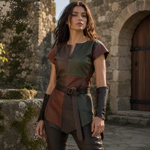 May include: A short-sleeved tunic crafted from panels of brown and dark green leather, featuring a notched neckline and a matching leather belt with a metal buckle. The garment is paired with dark leather arm bracers and dark fitted trousers.