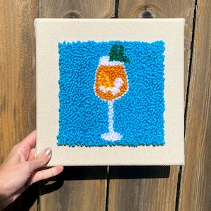 May include: A blue and white punch needle artwork of a glass of orange juice with a green garnish on a white canvas.