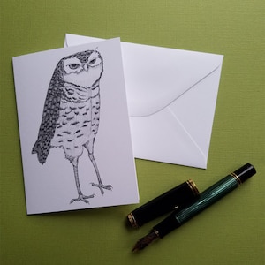 May include: A black and white illustration of an owl with a serious expression on a white card. The card is sitting on a green surface with a black and green pen next to it.