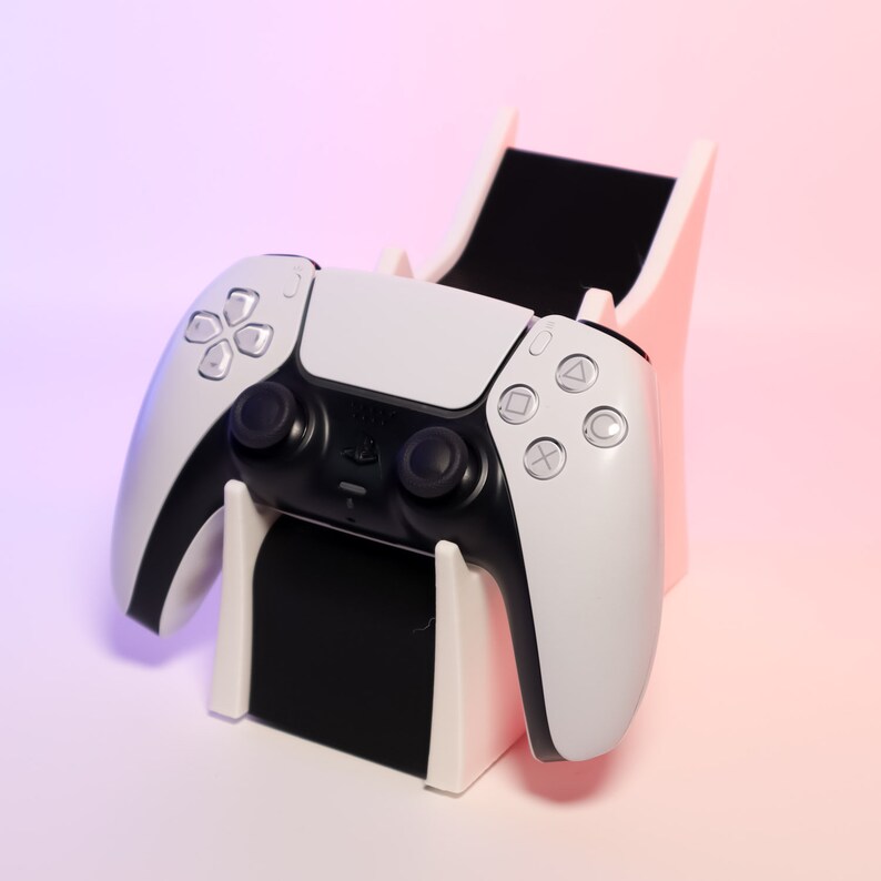 Op de afbeelding: Een witte en zwarte videogamecontroller staat in een roze en witte standaard. De controller heeft knoppen en een touchpad.
