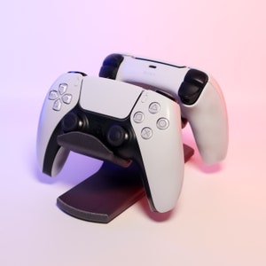 May include: Two white wireless game controllers with black buttons and a black charging stand. The controllers are on a white surface with a pink and blue gradient background.