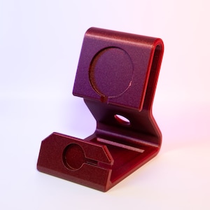 Puede incluir: Un soporte para teléfono de color rojo metálico con un acabado texturizado. El soporte tiene un diseño curvo con un recorte circular en la parte superior y una ranura en la base. Diseñado para sostener un teléfono o dispositivo similar.