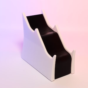 Puede incluir: Soporte de plástico blanco con un estante negro inclinado de varios niveles. El soporte está diseñado para contener objetos como libros u otros objetos decorativos.