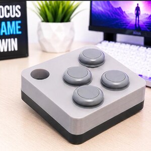 May include: A square-shaped macro keypad controller featuring a light gray top housing and a black base. The device includes four circular gray push buttons arranged in a diamond pattern and one circular cutout on the top left corner surface.