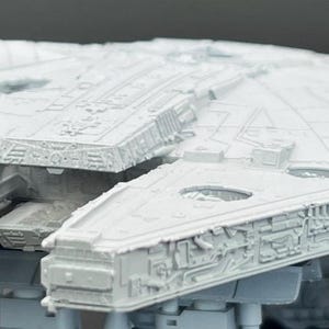 May include: A detailed, light gray model of the Millennium Falcon starship. The model features intricate detailing on the exterior, including panels, turrets, and a cockpit. The ship is positioned on a stand, showcasing its iconic design.