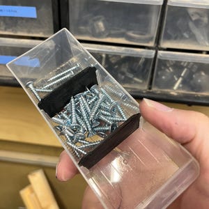 May include: A clear plastic container filled with silver screws. The container is held in a hand, and the screws are neatly organized inside. The background shows other storage containers.