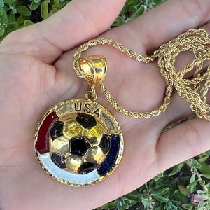 May include: A gold-toned rope chain necklace featuring a circular pendant with a central soccer ball design. The pendant includes the text USA at the top, with red, white, and blue enamel accents surrounding the soccer ball on a textured gold border.