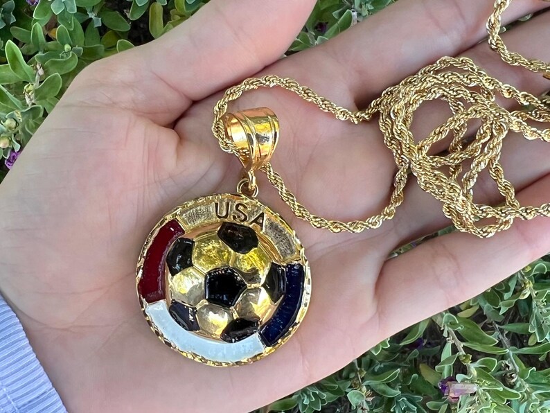 May include: A gold-toned rope chain necklace featuring a circular pendant with a central soccer ball design. The pendant includes the text USA at the top, with red, white, and blue enamel accents surrounding the soccer ball on a textured gold border.