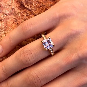 May include: A gold ring featuring a square, light purple gemstone. The ring's band is also gold and has small, decorative details. The ring is on a person's finger.