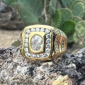 May include: A gold-toned ring with a large, oval-shaped, clear stone at the center, surrounded by smaller, clear stones. The ring has a square face with decorative detailing on the sides. The ring is set on a textured, gray rock.