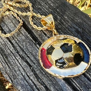 May include: A gold-tone pendant necklace featuring a central soccer ball design with black and gold panels. The circular pendant is accented with red, white, and blue enamel details along the outer rim and hangs from a twisted gold-tone rope chain.