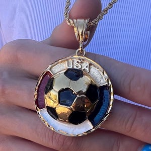 Pode incluir: Um pingente circular dourado em uma corrente de corda com uma bola de futebol tridimensional central com painéis pretos e dourados. A borda é acentuada com seções de esmalte vermelho, branco e azul e o texto USA está gravado no topo.