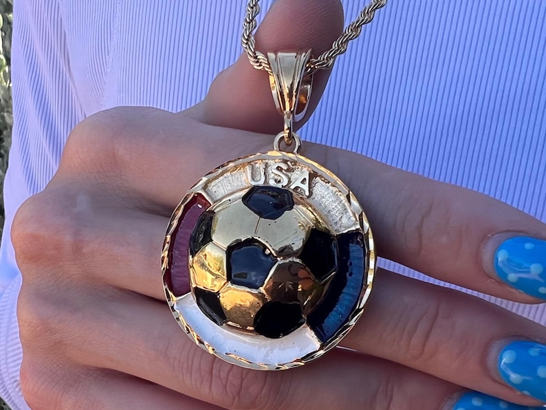 May include: A gold-toned circular pendant on a rope chain featuring a central three-dimensional soccer ball with black and gold panels. The border is accented with red, white, and blue enamel sections and the text USA is engraved at the top.