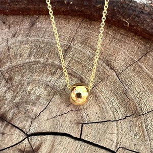 May include: A gold-colored necklace with a small, round pendant. The pendant is a shiny sphere, and the chain is delicate. The necklace is displayed on a textured, brown wooden surface.