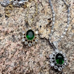 Puede incluir: Dos collares con colgantes de color verde esmeralda. Uno tiene una cadena y colgante de oro con un halo de piedras transparentes. El otro tiene una cadena y colgante de plata con un diseño similar.