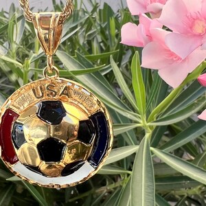 May include: A gold-tone circular pendant featuring a central soccer ball design with black and gold panels. The border includes the text USA at the top, with red, white, and blue enamel accents surrounding the soccer ball. It hangs from a gold rope chain.
