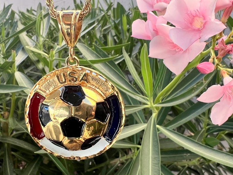 May include: A gold-tone circular pendant featuring a central soccer ball design with black and gold panels. The border includes the text USA at the top, with red, white, and blue enamel accents surrounding the soccer ball. It hangs from a gold rope chain.