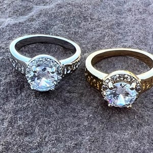 May include: Two rings, one silver-toned and one gold-toned, each featuring a large, clear, round gemstone surrounded by smaller stones. The bands have decorative detailing. The rings are displayed on a textured, gray surface.