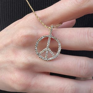 May include: A gold necklace with a peace symbol pendant. The pendant is set with small, clear gemstones, giving a sparkling appearance. The necklace is suspended from a fine gold chain.