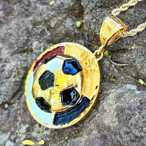May include: A gold-toned circular pendant featuring a raised soccer ball design with black enamel accents and the letters USA embossed on the side. The pendant hangs from a gold-toned rope chain, showcasing a polished metallic finish throughout.
