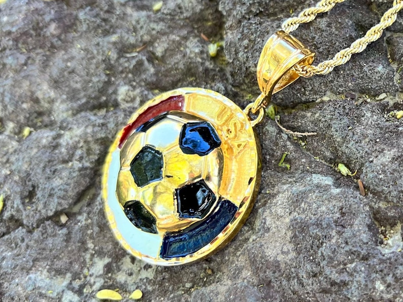 May include: A gold-toned circular pendant featuring a raised soccer ball design with black enamel accents and the letters USA embossed on the side. The pendant hangs from a gold-toned rope chain, showcasing a polished metallic finish throughout.