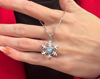 Op de afbeelding: Een zilveren ketting met een sneeuwvlokhanger met een lichtblauwe edelsteen in het midden. De hanger hangt aan een fijne ketting. De ketting wordt getoond tegen een rode en zwarte achtergrond.