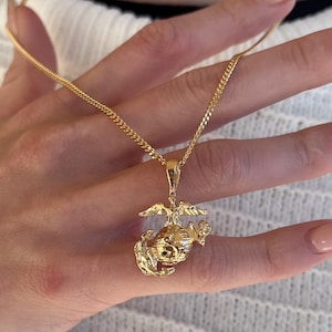 May include: A gold-coloured necklace with a pendant depicting an eagle, globe, and anchor emblem. The chain is a fine curb link design. The pendant and chain are shown against a plain backdrop.
