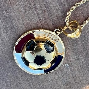 May include: A gold-tone metal pendant necklace featuring a circular design with a central soccer ball motif. The border includes red, white, and blue enamel accents and the text USA engraved at the top. The pendant hangs from a twisted rope chain.