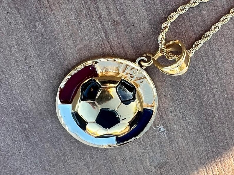May include: A gold-tone metal pendant necklace featuring a circular design with a central soccer ball motif. The border includes red, white, and blue enamel accents and the text USA engraved at the top. The pendant hangs from a twisted rope chain.
