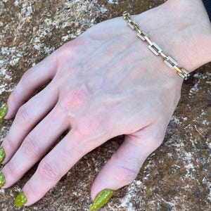 May include: A gold-toned link bracelet featuring rectangular links, each accented with small, clear, sparkling stones. The bracelet is worn on a wrist, showcasing the repeating geometric pattern of the metal links and the embedded stones.