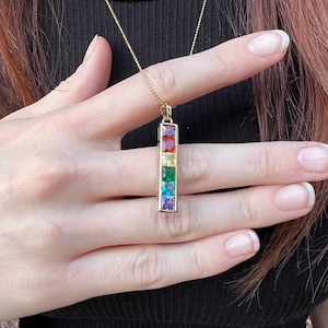 May include: A gold-colored necklace with a pendant featuring a vertical row of colorful rectangular gemstones. The gemstones are red, yellow, green, blue, and purple. The necklace is held up by a person wearing a black top.