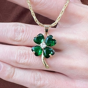Op de afbeelding: Een gouden ketting met een vierbladige klaver hanger. De klaver is gemaakt van groene edelstenen en heeft een kleine, heldere edelsteen in het midden.