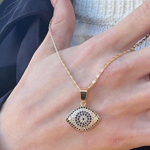 May include: A gold-colored necklace with a pendant shaped like an eye. The eye pendant is detailed with black and white gemstones. The necklace is worn on a person's neck.