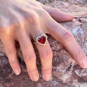 May include: A gold ring featuring a large red heart-shaped gemstone surrounded by smaller clear gemstones.