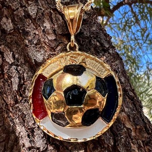 May include: A gold-toned circular pendant featuring a central soccer ball design with black and gold panels. The outer rim displays the text USA at the top, with red, white, and blue enamel accents surrounding the soccer ball. It includes a gold bail.