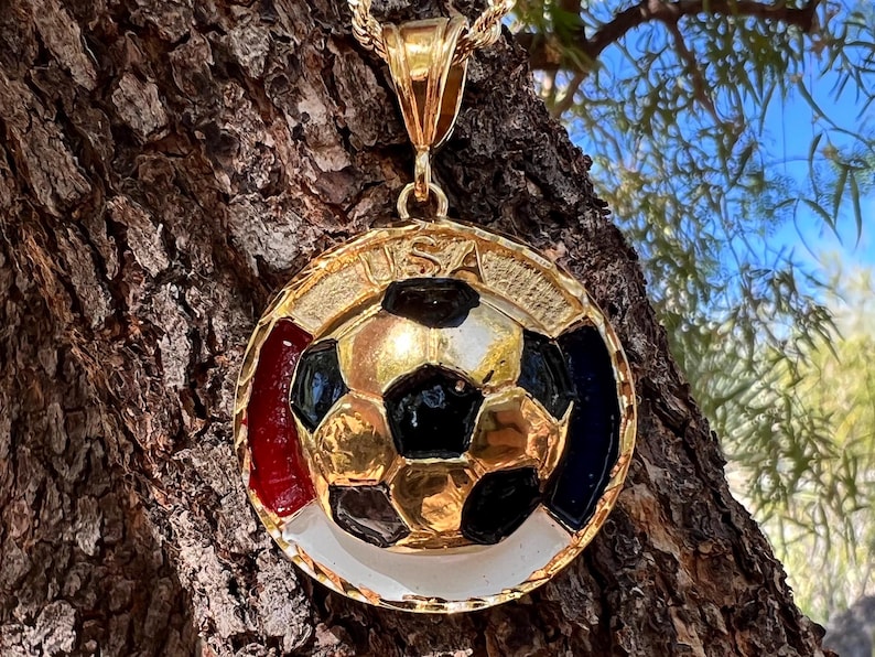 May include: A gold-toned circular pendant featuring a central soccer ball design with black and gold panels. The outer rim displays the text USA at the top, with red, white, and blue enamel accents surrounding the soccer ball. It includes a gold bail.