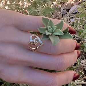 May include: A gold-toned ring featuring a heart-shaped outline with a stylized letter S integrated into the center. The upper curve of the heart is embellished with a row of small, clear, sparkling stones set along the metal band.