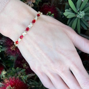 May include: A gold bracelet with oval-shaped red gemstones and small clear stones. The bracelet is on a wrist, surrounded by green leaves and red flowers. The bracelet's design features a repeating pattern of red and clear stones.