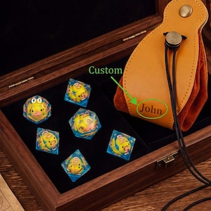 May include: A set of six clear polyhedral dice with yellow rubber ducks inside, displayed in a dark wooden box alongside a tan leather drawstring pouch featuring the custom text John embossed on the front surface of the leather material.